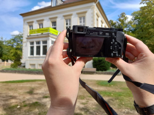 Zwei Hänge halten eine Fotokamera hoch. Im Hintergrund ist ein Haus.