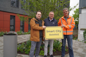 Jens Edler, Marcus Lufen und Stefan Meyer halten ein Schild mit der Aufschrift Smart City in Händen. Neben ihnen ist einer der Abfallbehälter zu sehen